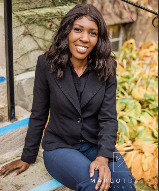 Headshot of Alexis Whitehorn wearing a black blazer jacket, jeans and sitting outside of a building beside green and orange plants