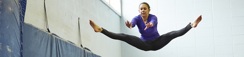 Women wearing a purple t-shirt and leggings jumping on a trampoline while doing side splits