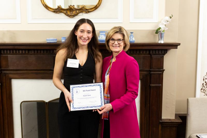 Hannah Chown poses with Dean Gretchen Kerr, holding her Hill-Powell Award in front of the Arts & Letters club fireplace