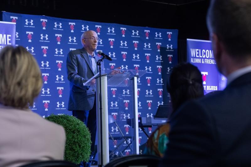 KPE alumni and donor Michael Guinness stands in front of a branded display while addressing the attendees of the 2025 Celebration of Impact