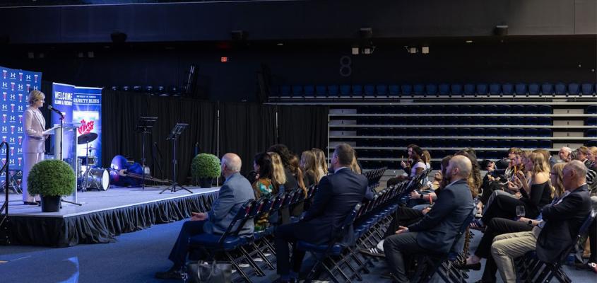 KPE Dean Gretchen Kerr addresses an audience of donors and alumni, standing on a stage in front of KPE branding in the Kimel Family Field House