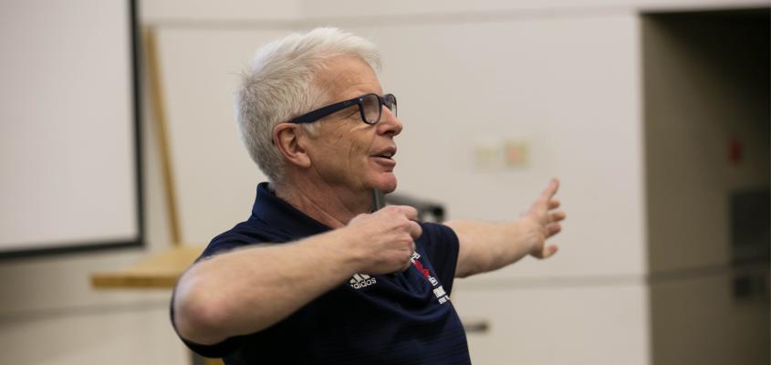 Doug Richards gestures while giving a lecture wearing a blue polo with the U of T Varsity Blues leaf logo