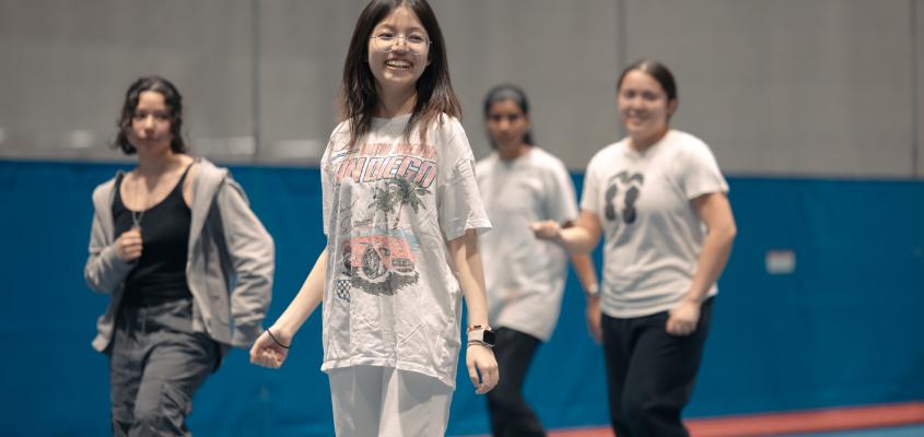 A group of smiling university students with a girl in focus in the centre. She is fair with dark brown hair wearing round silver glasses and a baggy tshirt, smiling big looking to the side.