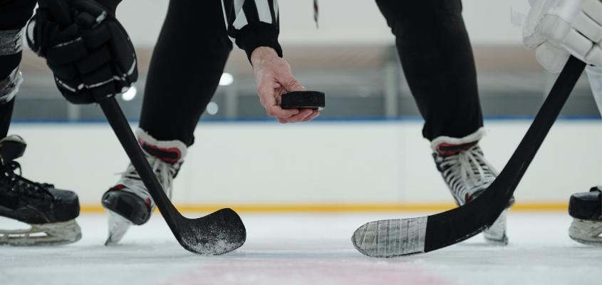 Hand of referee holding puck over ice rink with two players with sticks standing around and waiting for moment to shoot it