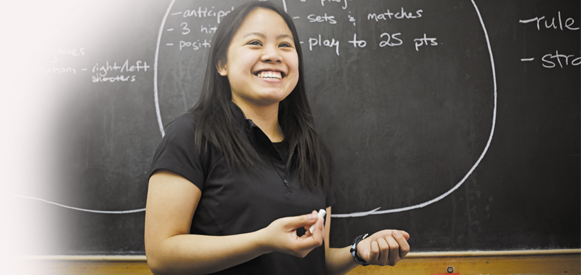 a smiling female undergraduate student in front of a chalkboard explaining game rules