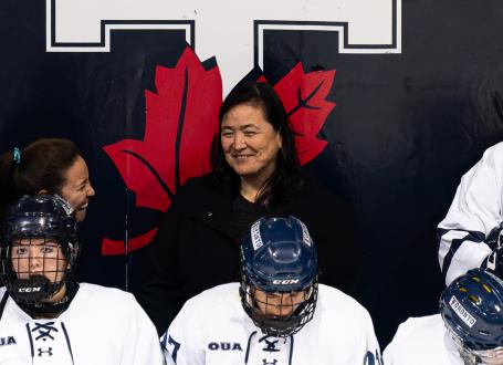 U of T Varsity Blues women's hockey head coach Vicky Sunohara smiles during a game behind players seated on the bench.