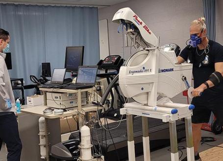 A researcher stands in front of a computer console while a study participant uses an arm bike while wearing an array of testing equipment attached to his face.